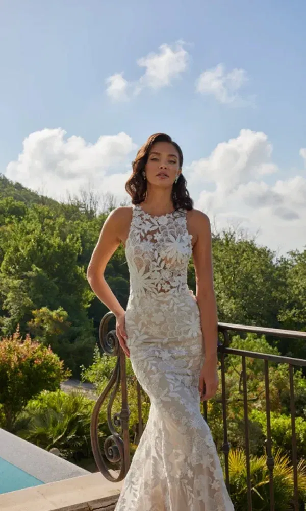 Stilvolle Braut mit enganliegendem Spitzenkleid lehnt an schmiedeeisernem Geländer mit blauem Himmel im Hintergrund.