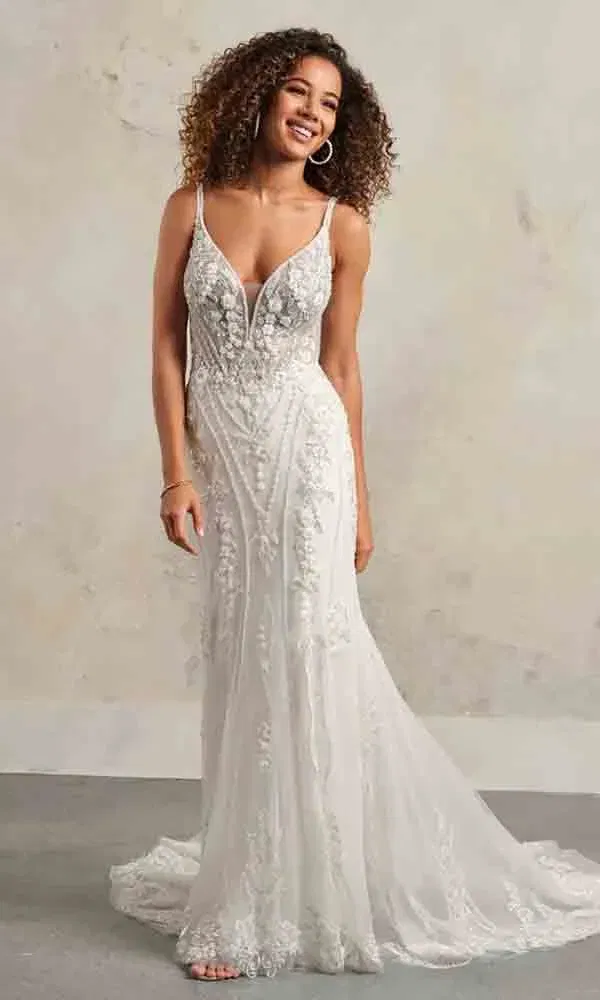 Glückliche Braut in figurbetontem Brautkleid mit floralem Muster und transparentem Ausschnitt, lächelt vor hellem Hintergrund.