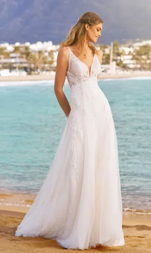 Eine romantische Braut im A-Linien-Brautkleid genießt den Ausblick am Strand mit Meer und Küstenstadt im Hintergrund.