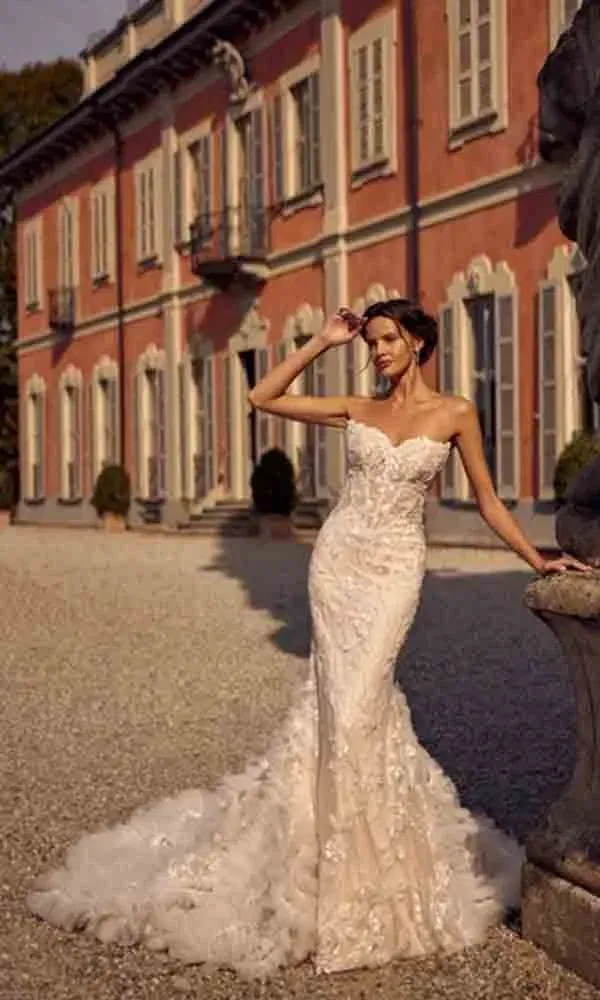 Strapless-Brautkleid mit Tüllschleppe und floralen Details – die Braut lehnt entspannt an einem Steinpfeiler vor einer herrschaftlichen Villa.