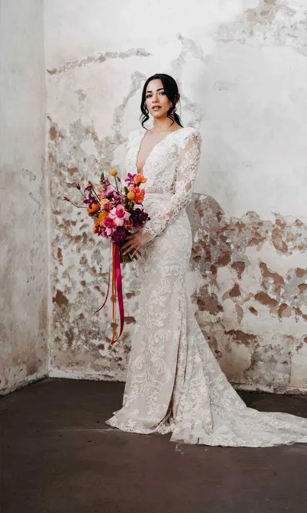 Moderne Braut in cremefarbenem Spitzenkleid mit V-Ausschnitt und farbenfrohem Strauß vor verwitterter Wand