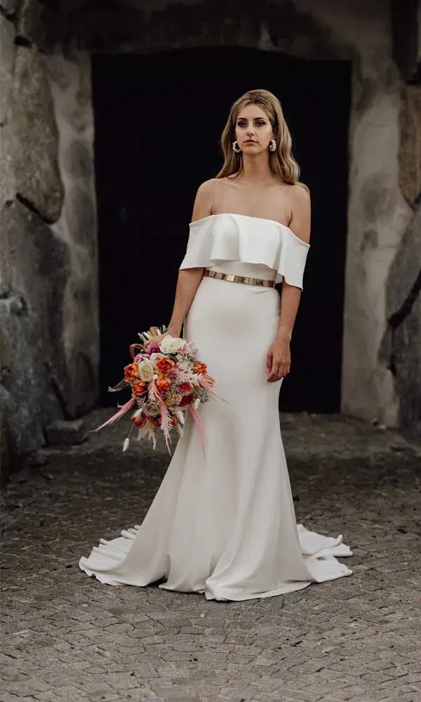 Elegante Frau im modernen Brautkleid mit Gürtel und sattem Blumenstrauß auf Kopfsteinpflaster vor dunklem Torbogen