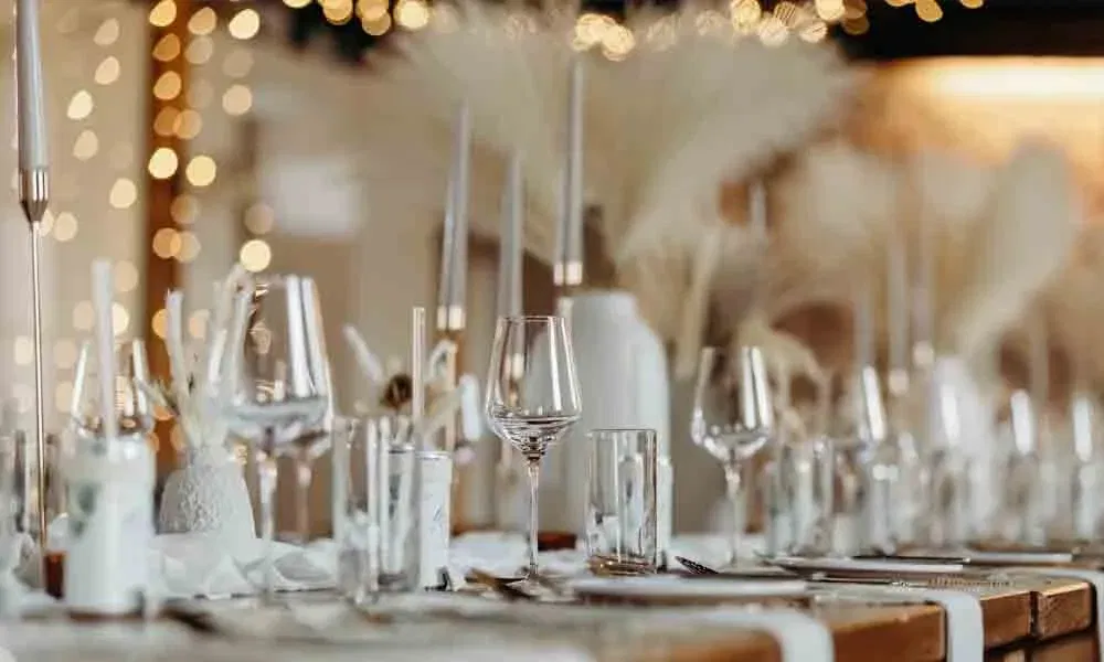 Stimmungsvolle Nahaufnahme eines gedeckten Holztisches bei einer Hochzeit, dekoriert mit Pampasgras, Windlichtern und Lichterketten im Hintergrund.
