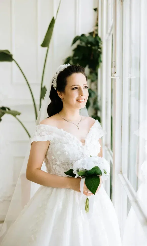 Romantischer Moment einer Braut mit Hochzeitsstrauß im Licht eines hellen Zimmers am Fenster.