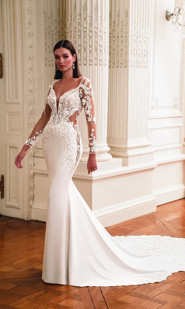 Stilvolle Braut in einem enganliegenden Kleid mit transparenten Cut-outs, langen bestickten Ärmeln und Schleppe, stehend in einem weißen, opulenten Raum mit historischen Details.