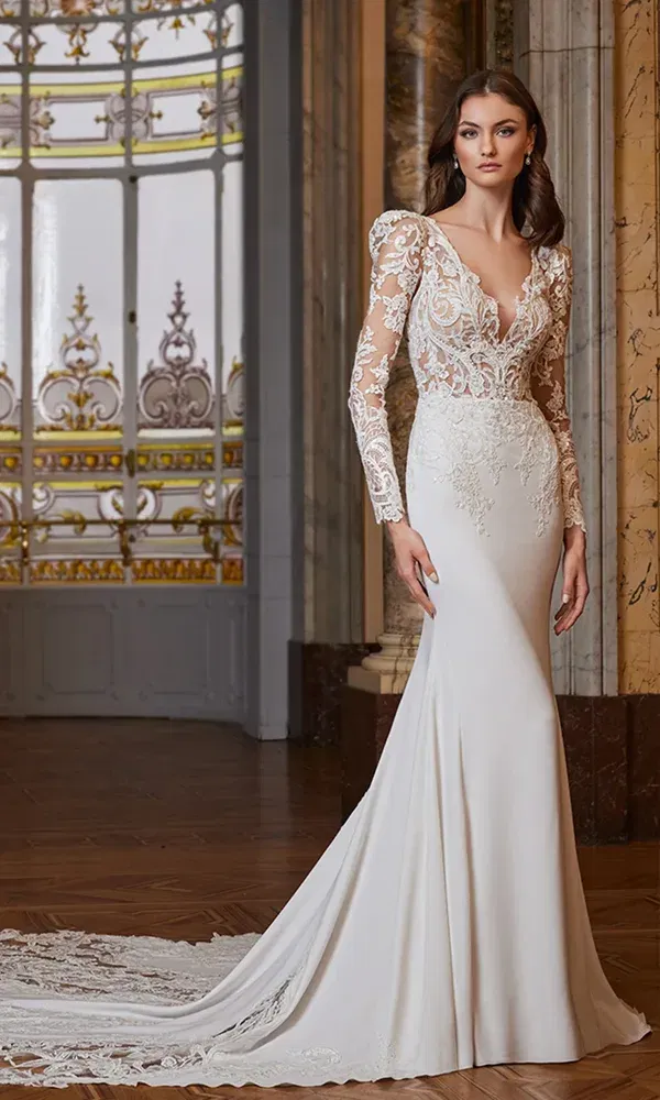 Model trägt ein romantisches Hochzeitskleid mit transparenter Spitzen-Oberpartie, langen Ärmeln und feiner Schleppe, posierend vor marmorierten Säulen und Glasornamenten.