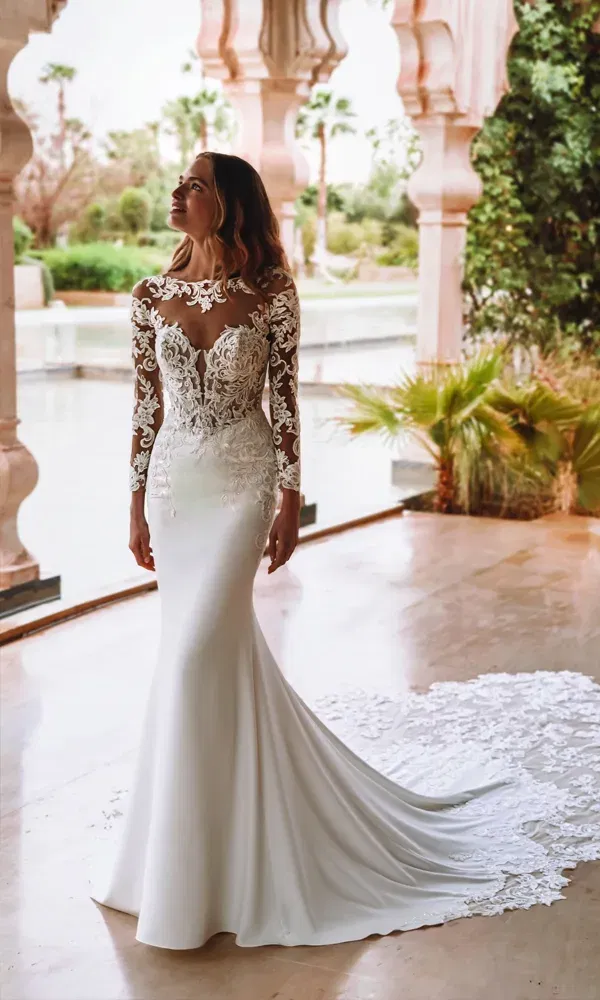 Fröhliche Braut trägt ein figurbetontes Hochzeitskleid mit Spitzenoberteil und geschlossener Halslinie, stehend in einem orientalisch inspirierten Außenbereich mit Wasserblick.