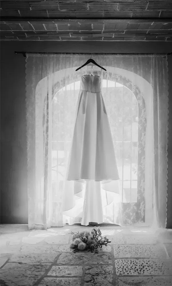 Schlichtes, modernes Brautkleid vor einer Fensterkulisse aus Spitze – liebevoll inszenierte Vorbereitungsaufnahme in Schwarz-Weiß.