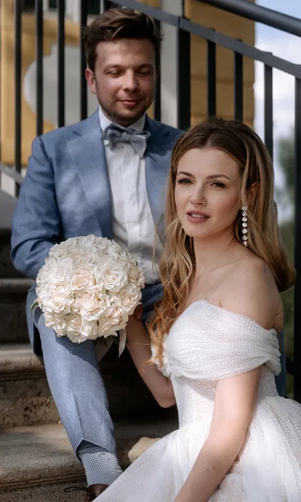 Sitzendes Brautpaar auf einer Treppe, die Braut trägt ein schulterfreies Kleid und hält einen großen Strauß weißer Rosen.