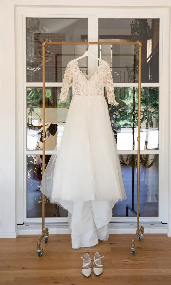 Elegantes Brautkleid mit Spitze und Tüll hängt an einem goldenen Kleiderständer, darunter passende Brautschuhe.