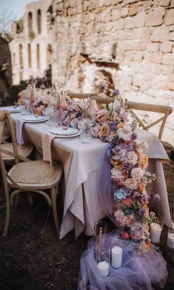 Romantisch dekorierter Hochzeitstisch mit pastellfarbener Blumengirlande vor rustikaler Steinwand im Freien.
