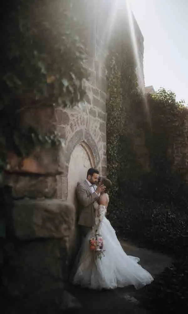Zärtliche Szene eines Brautpaares im Sonnenstrahl vor romantischer Kulisse mit historischem Flair.