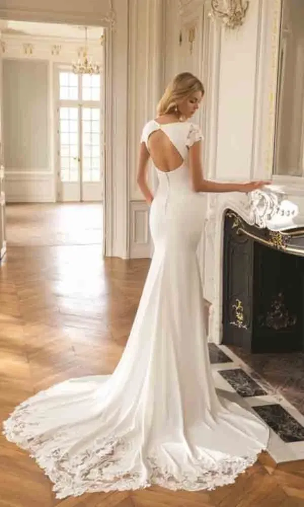 Minimalistisches Brautkleid mit rundem Rückenausschnitt und romantischer Schleppe, inszeniert in einem lichtdurchfluteten Saal.