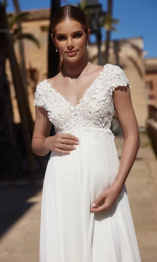 Im Freien aufgenommene Szene mit einer schwangeren Frau im eleganten Hochzeitskleid mit floralen Details.