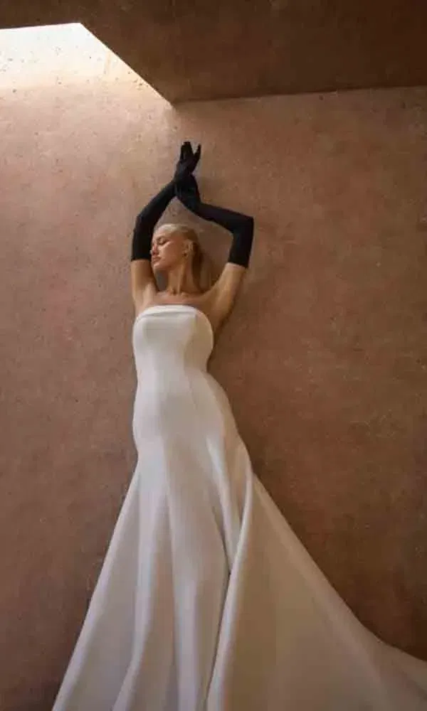 Schlichtes Designerbrautkleid mit langer Schleppe und schwarzen Accessoires, elegant fotografiert vor Terrakottawand.