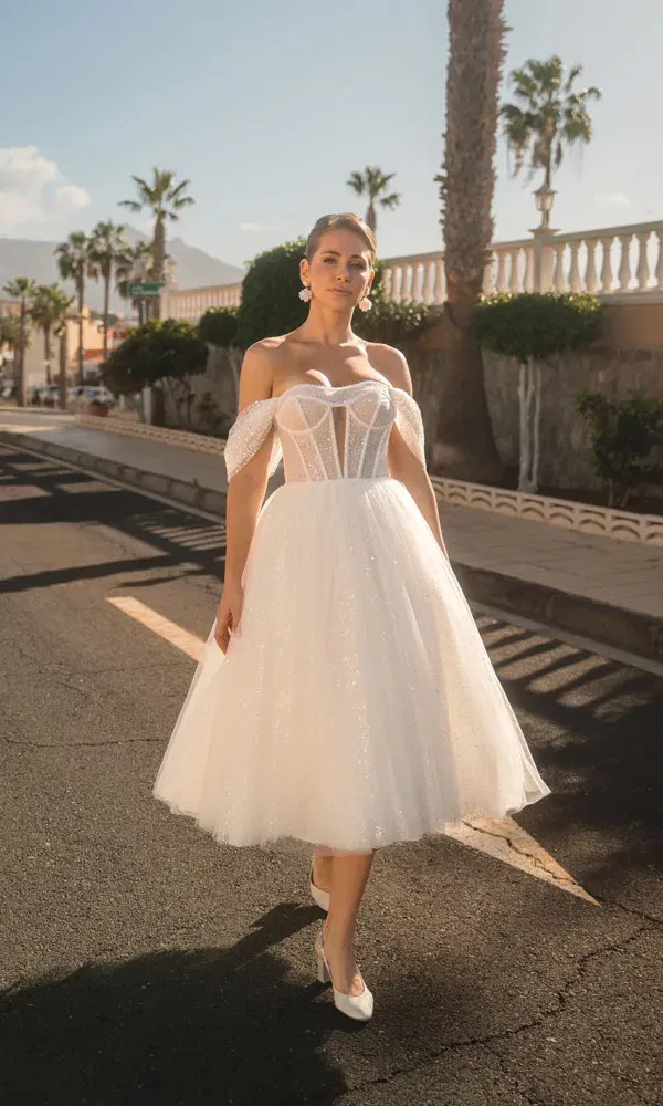Romantischer Bridal-Look: Braut im glitzernden Midikleid mit offener Schulterpartie geht stilvoll auf einer warmen, südlichen Straße entlang.