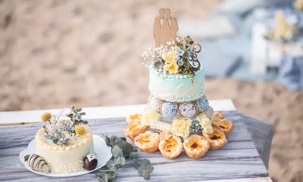 Strand-Hochzeitstorte mit zartblauer Deko, Blüten, Topper für zwei Bräutigame und Mini-Pastéis de Nata, daneben eine zweite Torte mit romantischer Verzierung.