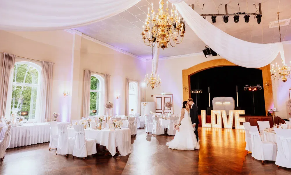 Ein festlich geschmückter Hochzeitssaal mit hoher Decke, Lichterketten und einem Brautpaar in der Mitte vor einem beleuchteten LOVE-Schriftzug.