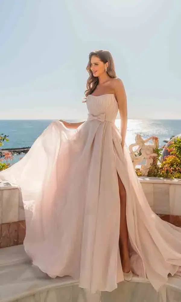 Strahlende Frau in luftigem, trägerlosen Brautkleid mit ausgestelltem Rock und Beinschlitz vor maritimem Hintergrund.