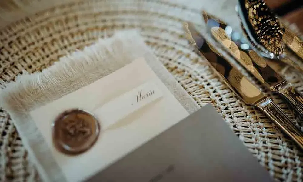 Boho-inspirierte Papeterie bei einer Hochzeit: Namenskärtchen mit Siegel auf strukturiertem Stoff, daneben silbernes Besteck auf geflochtenem Platzset.