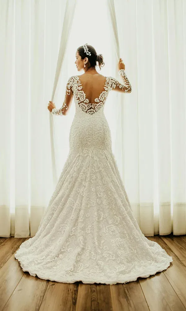 Frau in Brautkleid mit transparenter Spitzenrückseite zieht die Vorhänge zur Seite.