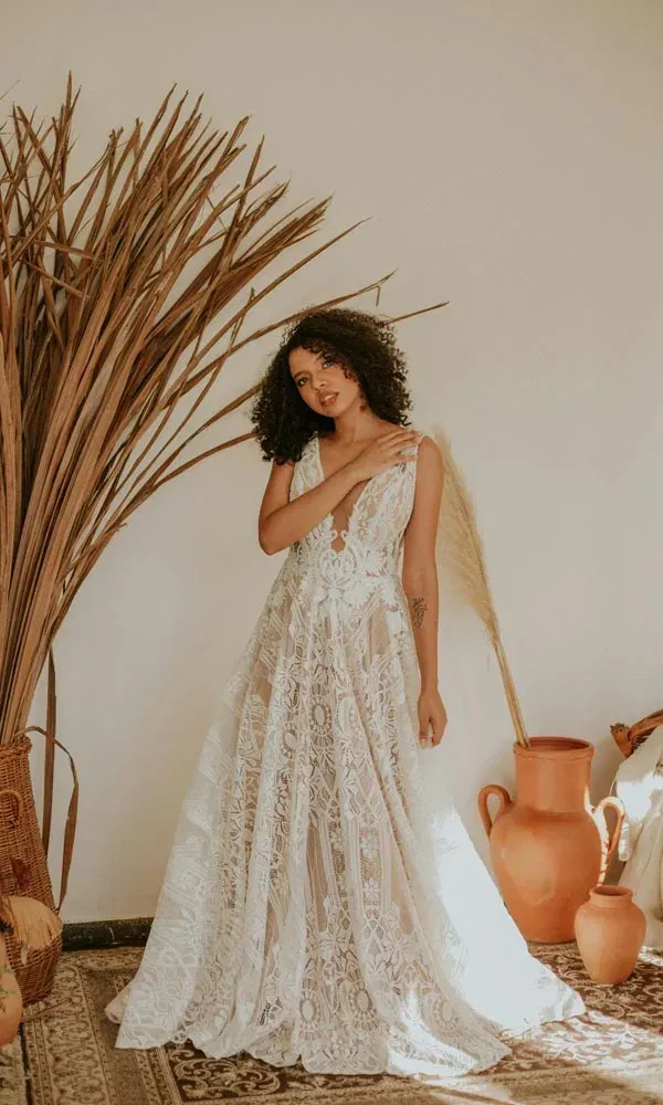 Eine elegante Braut mit Locken trägt ein durchsichtiges Spitzenkleid im Boho-Look und steht neben getrocknetem Palmblatt.