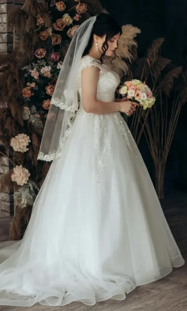 Elegante Braut mit Lockenfrisur hält einen Strauß Rosen und trägt ein besticktes Hochzeitskleid.