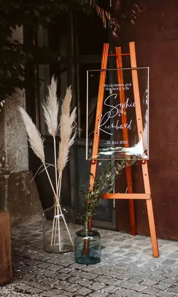 Dekorative Willkommensstation bei einer Sommerhochzeit mit Acrylschild, Holzstaffelei und natürlicher Blumendeko im Boho-Stil.