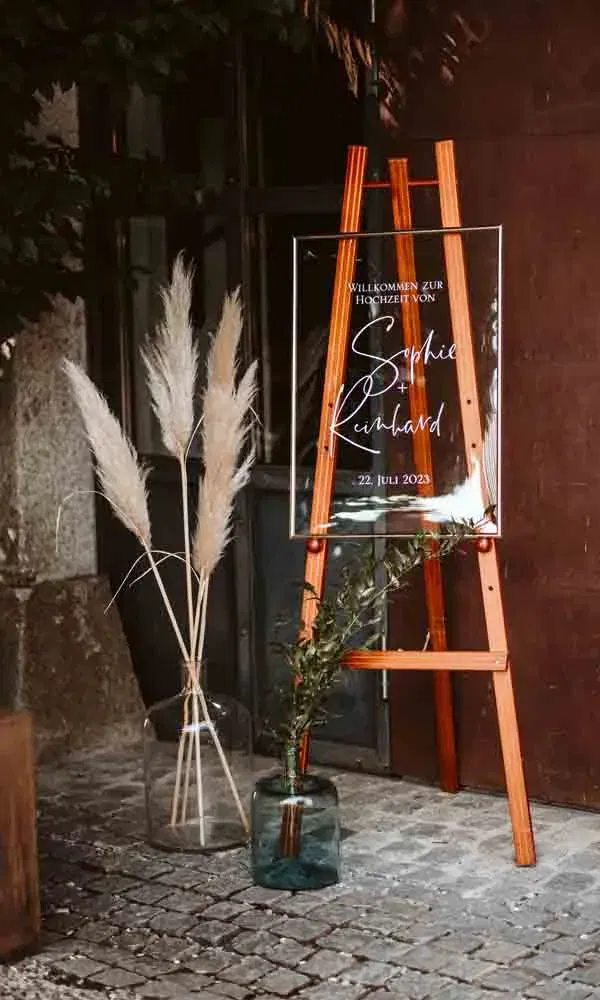 Dekorative Willkommensstation bei einer Sommerhochzeit mit Acrylschild, Holzstaffelei und natürlicher Blumendeko im Boho-Stil.