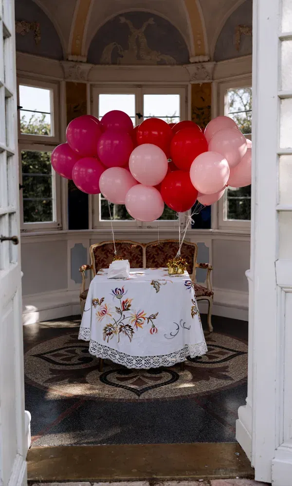 Dekorativer Hochzeitstisch mit Blumenmuster, Goldgeschenk und rosafarbenen Luftballons in einem historischen Pavillon.