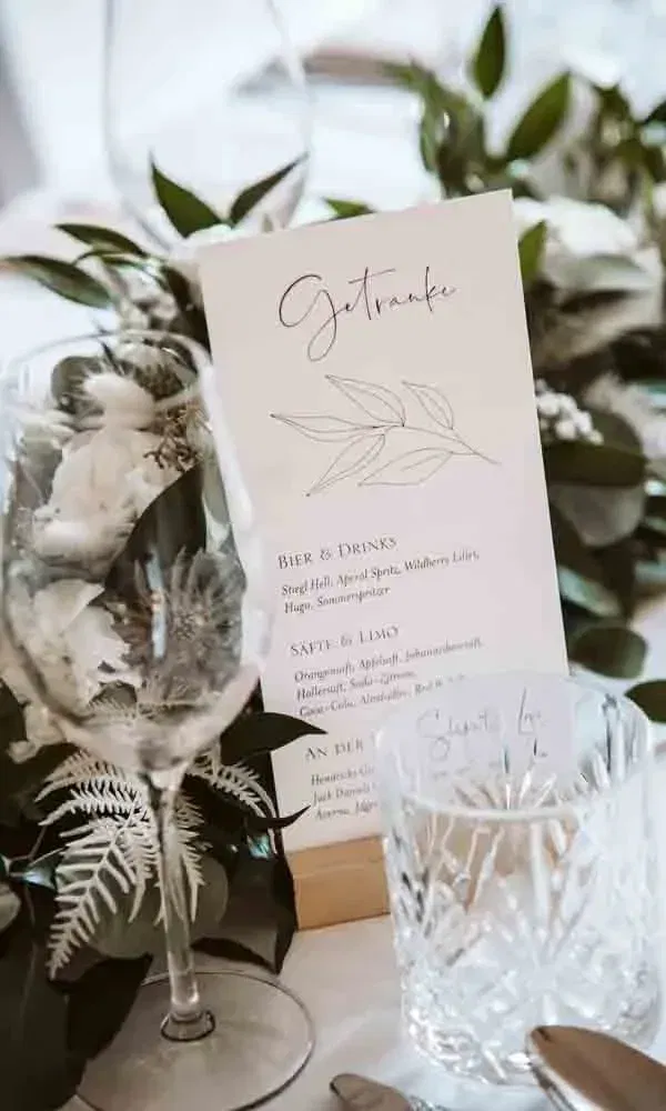 Detailaufnahme einer Hochzeits-Getränkekarte mit filigraner Typografie und floraler Tischdekoration im Hintergrund.
