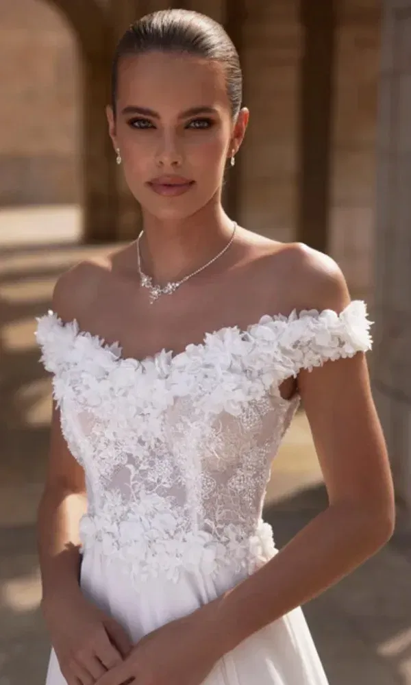 Braut in einem floralen, schulterfreien Hochzeitskleid posiert selbstbewusst vor einer mediterranen Steinfassade.