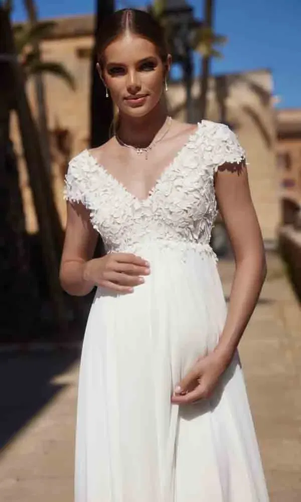 Junge Frau in einem weißen Schwangerschaftsbrautkleid mit Blütendetails und sommerlicher Kulisse im Hintergrund.