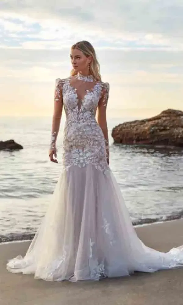 Braut in figurbetontem Tüllkleid mit tiefem Dekolleté und langen Spitzenärmeln am Wasser, umgeben von Felsen.