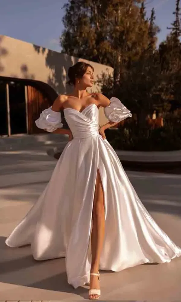 Fashion-forward Braut in weißem Satin-Hochzeitskleid mit Puffärmeln und sexy Beinschlitz auf urbaner Terrasse.