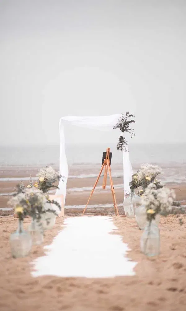 Minimalistischer Hochzeitsgang im Sand mit weißen Stoffen, einem Traubogen und floraler Deko – bereit für eine freie Trauung am Wasser.