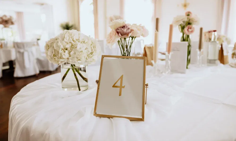 Elegante Hochzeitsdekoration auf Tisch Nummer 4 mit weißen Blüten, rosafarbenen Akzenten und fein drapierter Tischdecke.