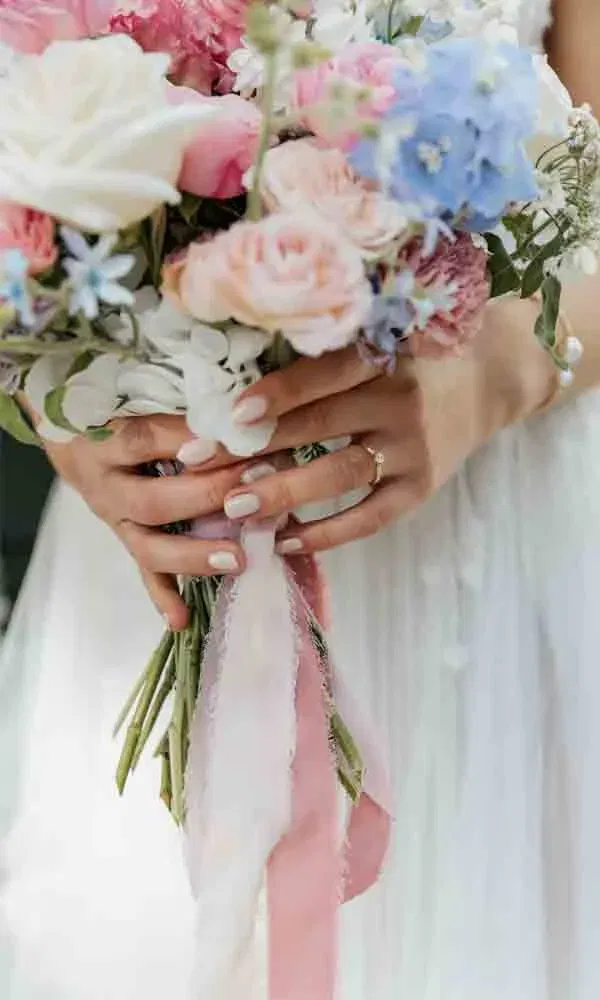 Pastellfarbener Blumenstrauß mit Schleifen in Rosé, liebevoll von der Braut umklammert – Fokus auf Blüten und Fingernägeln.