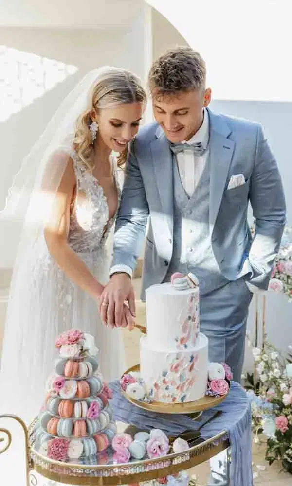 Hochzeitstortenmoment mit stilvollem Brautpaar, umgeben von liebevoll dekorierten Macarons und floralen Details in Pastellfarben.