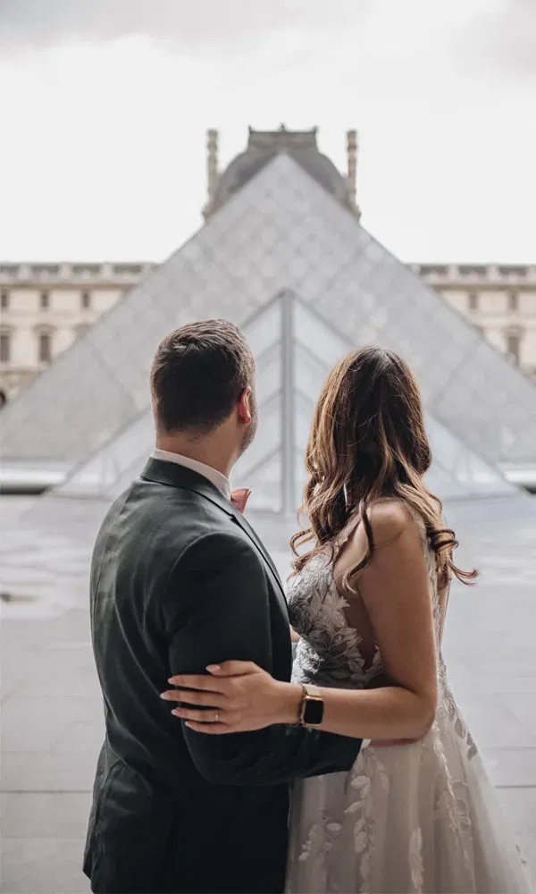 Hochzeitspaar steht Arm in Arm vor der Louvre-Pyramide und genießt den Moment in der Stadt der Liebe.
