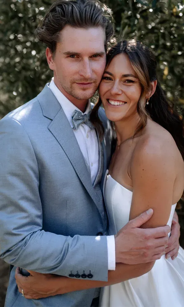 Zärtliche Umarmung des Brautpaars mit Blickkontakt zur Kamera – inniger Porträtmoment bei Tageslicht im Grünen.