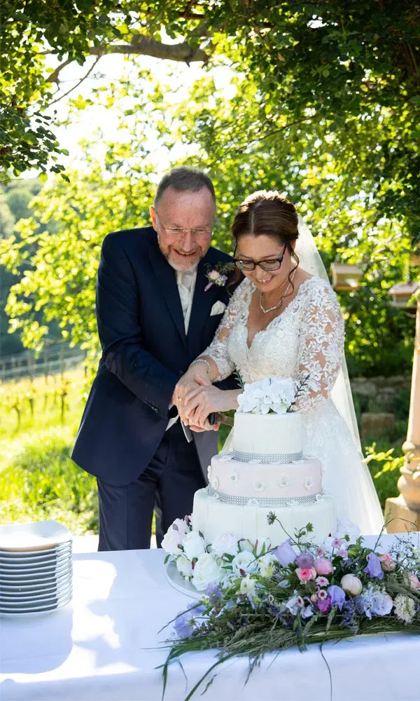 Braut und Bräutigam schneiden gemeinsam ihre dreistöckige Hochzeitstorte im Garten an.