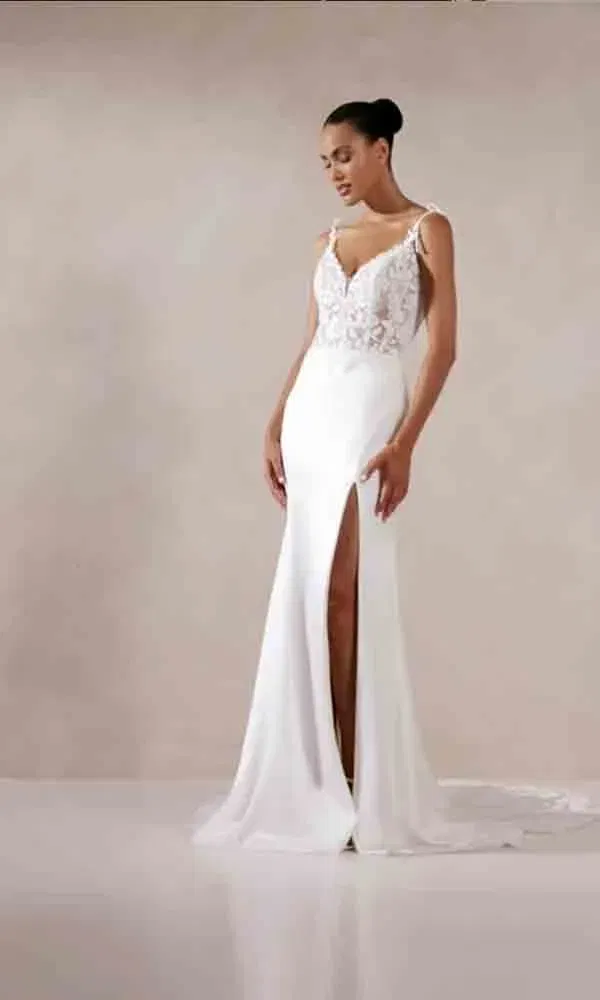 Schmal geschnittenes Hochzeitskleid mit filigranem Oberteil und sexy Beinschlitz, fotografiert vor einem sanft getönten Hintergrund in klassischer Pose.