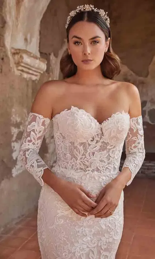 Körpernahes Off-Shoulder-Brautkleid mit detailreicher Spitze und floraler Stickerei vor rustikaler Steinwand.