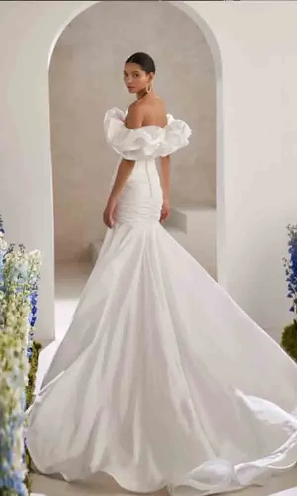 Figurbetonte Hochzeitsrobe mit strukturierter Schleppe und off-shoulder Volants, getragen von einer Braut im klassischen Setting mit floraler Dekoration.