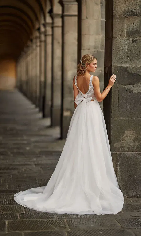 Elegantes Brautkleid mit tiefem Rückenausschnitt, Spitzen-Trägern und luftiger Schleppe – Braut lehnt an Steinmauer in gewölbtem Korridor.