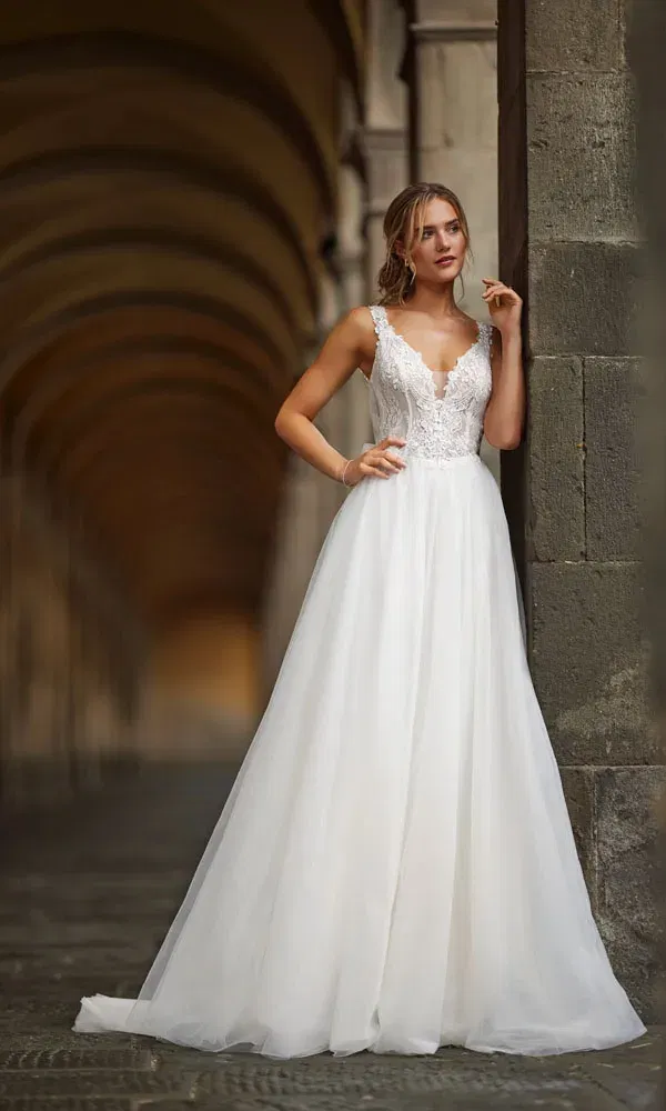 Verspielte Braut in Spitzenkleid mit transparentem Oberteil und voluminösem Rock – Szene in arkadenartiger Location mit sanftem Licht.