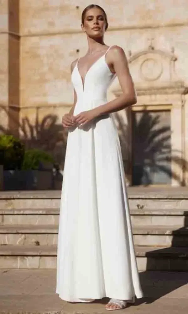 Moderne Brautmode in minimalistischem Stil – weißer Einteiler mit weitem Bein, präsentiert vor mediterranem Gebäude.