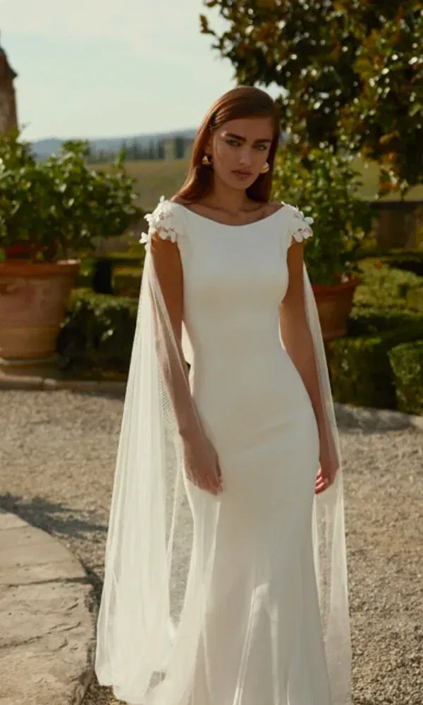 Moderne Braut in minimalistischem Kleid mit Capeärmeln, aufgenommen in einer mediterranen Parklandschaft.