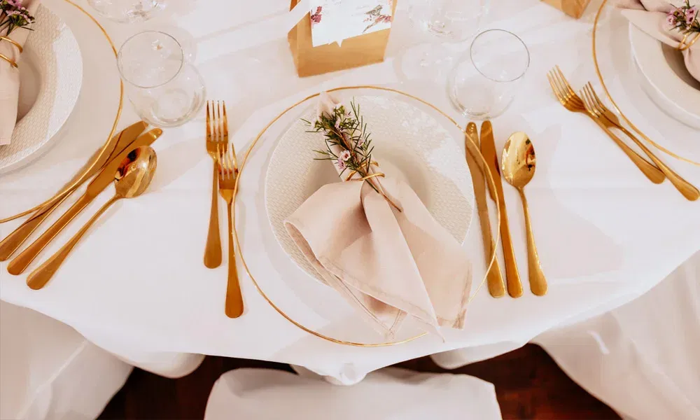 Detailaufnahme eines Hochzeitstisches mit edlem Goldbesteck, hellem Geschirr und Servietten, dekoriert mit frischen Zweigen und Blüten.